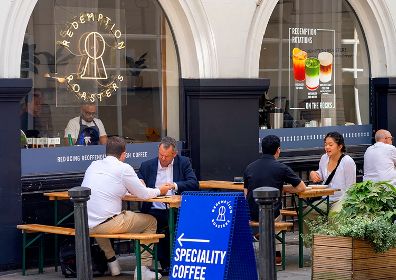 Redemption Roasters is a well-known coffee
chain located around London. Two people enjoy
coffee outside sat on tables in a relaxed setting.