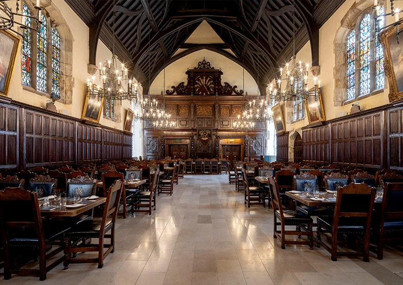 The Old Hall is a grand, historic dining hall with
high vaulted ceilings and dark wooden beams.
The room is lined with long wooden tables set
with silverware and glassware. Ornate
chandeliers hang from the ceiling, casting warm
light. Stained glass windows and large oil
paintings decorate the stone walls, and an
intricately carved wooden gallery and organ are
visible at the far end of the hall.
