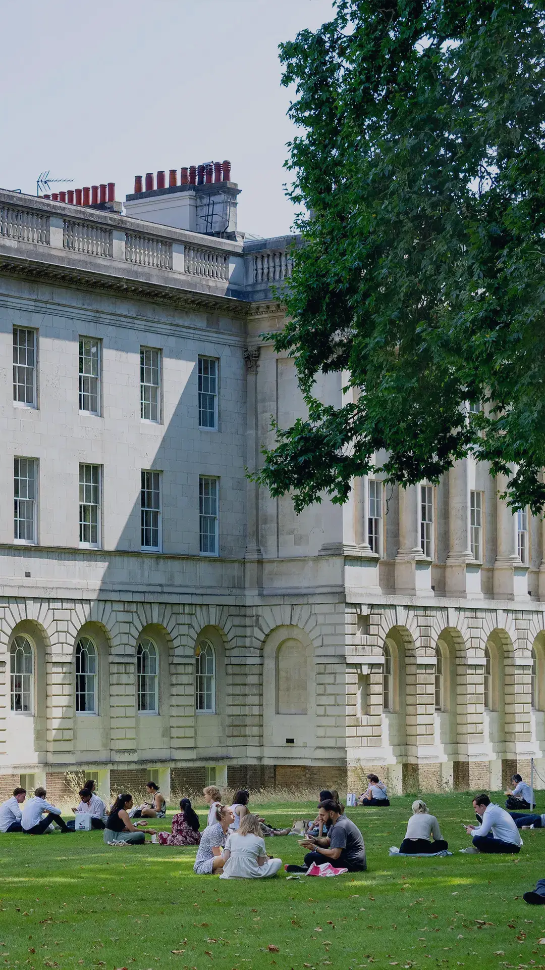 Stone building and walkways can be found on
Lincoln’s Inn Estate. People sit to eat lunch on
the well-maintained green lawn in front of the
buildings, there are multiple leafy trees that
provide shade.